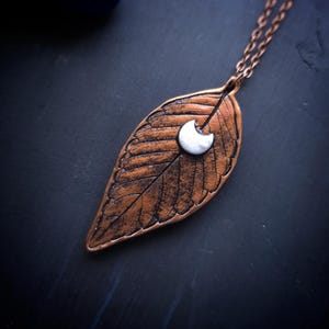 May include: A copper-colored leaf pendant necklace with a white crescent moon charm. The leaf pendant has detailed veining and is suspended from a delicate chain. The necklace is set against a dark background.