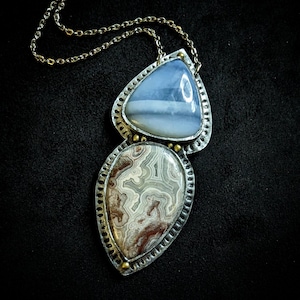 May include: A silver necklace with a two-toned agate pendant. The pendant is a teardrop shape with a light blue agate on top and a brown and white agate on the bottom. The necklace is made of a gold chain.