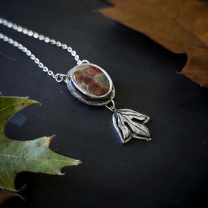 May include: A silver pendant necklace with an oval-shaped stone featuring brown and tan patterns. A silver leaf charm hangs below the stone. The necklace rests on a dark surface with green and brown leaves.