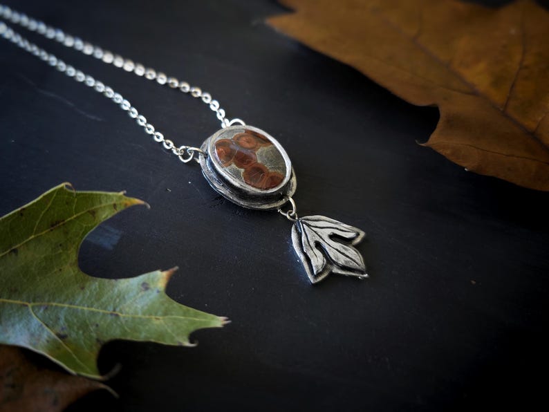 May include: A silver pendant necklace with an oval-shaped stone featuring brown and tan patterns. A silver leaf charm hangs below the stone. The necklace rests on a dark surface with green and brown leaves.