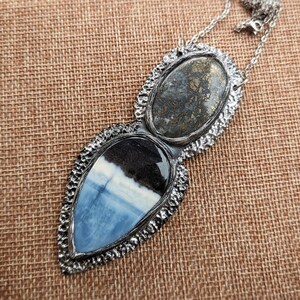 May include: A silver-toned pendant necklace featuring two stone-set elements. The top oval stone has a mottled gray appearance, while the lower, teardrop-shaped stone displays blue, white, and black patterns. The necklace is on a brown woven surface.