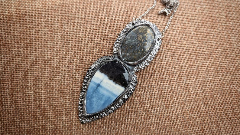 May include: A silver-toned pendant necklace featuring two stone-set elements. The top oval stone has a mottled gray appearance, while the lower, teardrop-shaped stone displays blue, white, and black patterns. The necklace is on a brown woven surface.