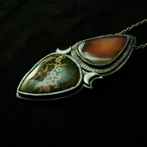 May include: A silver pendant necklace with two teardrop-shaped stones. The top stone is a light orange agate, and the bottom stone is a green and brown jasper with a speckled pattern.