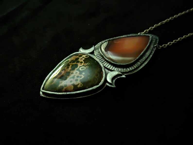 May include: A silver pendant necklace with two teardrop-shaped stones. The top stone is a light orange agate, and the bottom stone is a green and brown jasper with a speckled pattern.