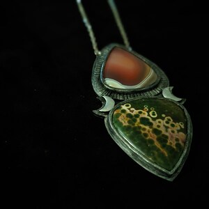 May include: A silver pendant necklace with two teardrop-shaped stones. The top stone is a red and white agate, and the bottom stone is a green and brown jasper. The pendant is surrounded by a silver crescent moon design.