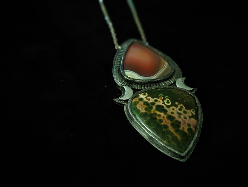 May include: A silver pendant necklace with two teardrop-shaped stones. The top stone is a red and white agate, and the bottom stone is a green and brown jasper. The pendant is surrounded by a silver crescent moon design.