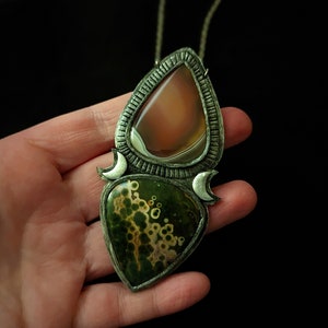 May include: A silver pendant necklace with two teardrop-shaped stones. The top stone is a light brown agate with white bands. The bottom stone is a green and brown moss agate with white spots. The pendant is surrounded by a silver frame with crescent moon shapes.