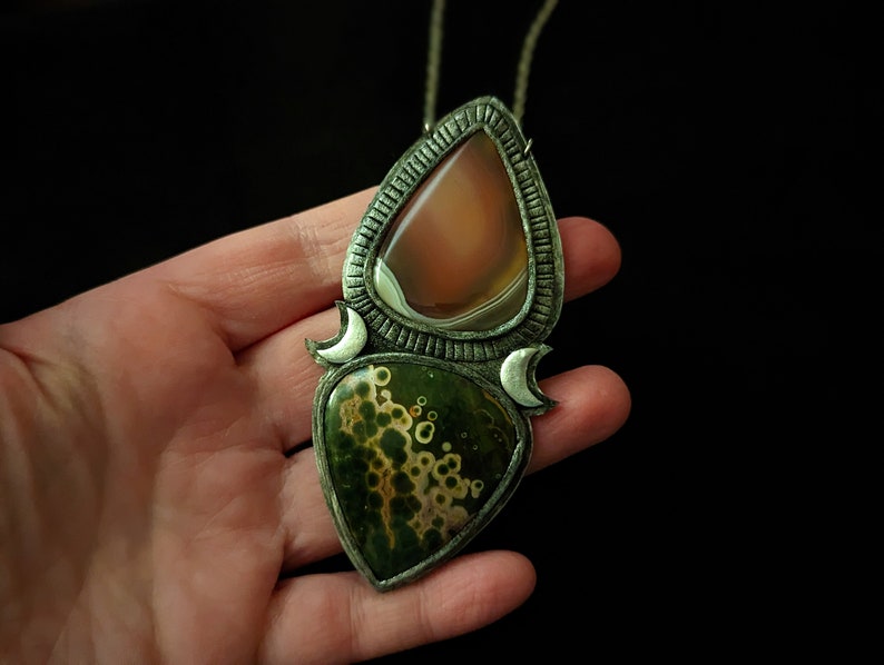 May include: A silver pendant necklace with two teardrop-shaped stones. The top stone is a light brown agate with white bands. The bottom stone is a green and brown moss agate with white spots. The pendant is surrounded by a silver frame with crescent moon shapes.