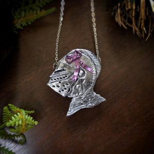 this image shows a hand sculpted knights helmet in silver color. the helmet is decorated with a pink bow