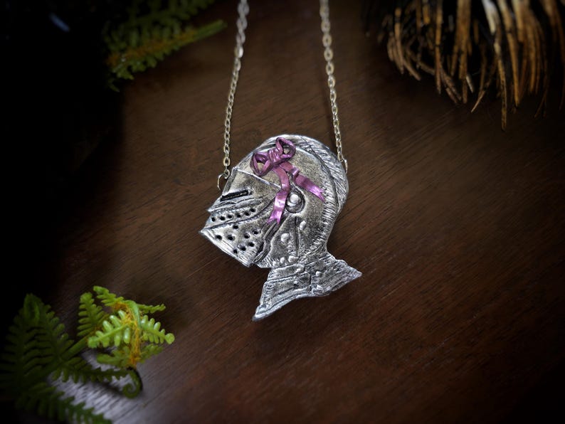 this image shows a hand sculpted knights helmet in silver color. the helmet is decorated with a pink bow
