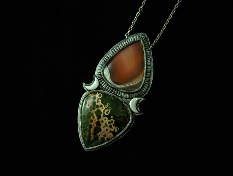 May include: A silver pendant necklace with two teardrop-shaped stones. The top stone is a reddish-brown agate, and the bottom stone is a green and white jasper. The pendant is accented with silver crescent moons.