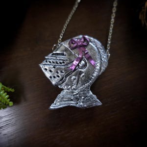 this image shows a hand sculpted knights helmet in silver color. the helmet is decorated with a pink bow