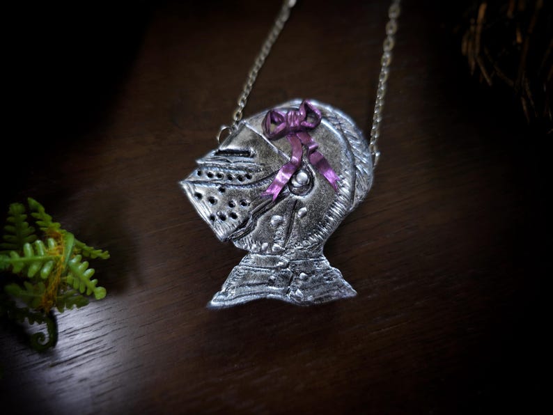 this image shows a hand sculpted knights helmet in silver color. the helmet is decorated with a pink bow