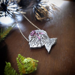 this image shows a hand sculpted knights helmet in silver color. the helmet is decorated with a pink bow