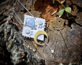 May include: A silver chain necklace with a silver and gold pendant featuring a crescent moon, stars, and a moonstone. The pendant is attached to a silver chain with a small bead at the end.