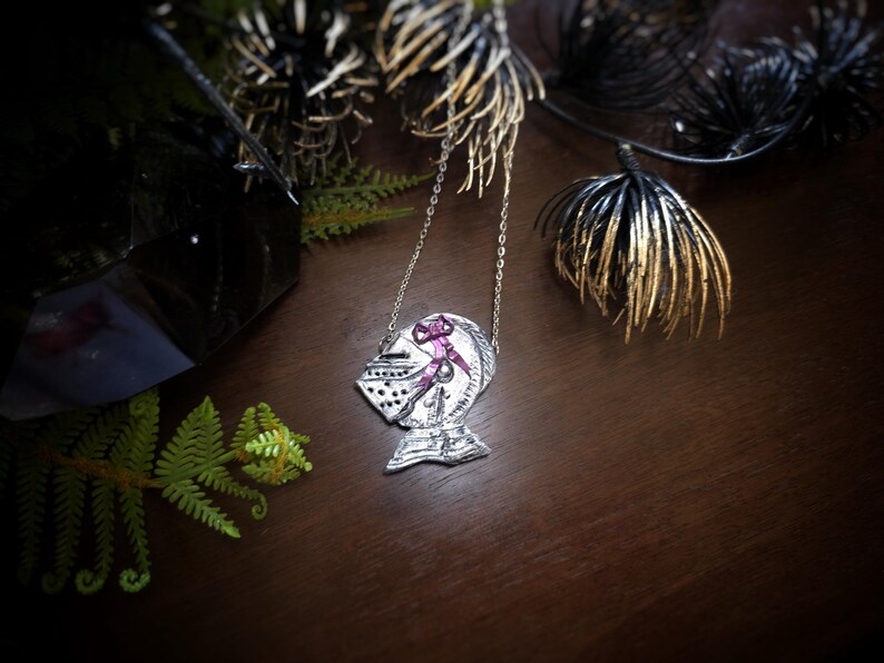this image shows a hand sculpted knights helmet in silver color. the helmet is decorated with a pink bow