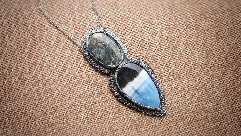 May include: A silver necklace with a pendant featuring two stone settings. The top stone is oval-shaped with gray and gold tones, while the bottom is a teardrop shape with blue, white, and black hues.