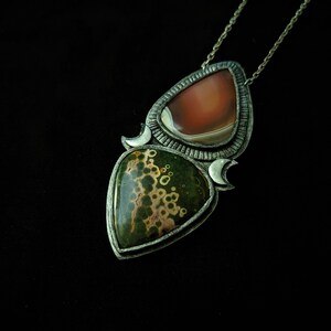 May include: A silver pendant necklace with two teardrop-shaped stones. The top stone is a light brown agate, and the bottom stone is a green and brown jasper with a speckled pattern.