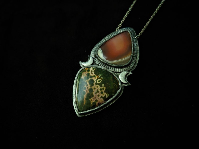 May include: A silver pendant necklace with two teardrop-shaped stones. The top stone is a light brown agate, and the bottom stone is a green and brown jasper with a speckled pattern.