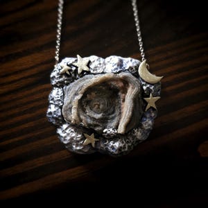 May include: A silver necklace with a pendant featuring a central, textured stone in shades of gray and beige. The pendant is surrounded by a silver cloud-like frame with small star and crescent moon accents. The chain is also silver.