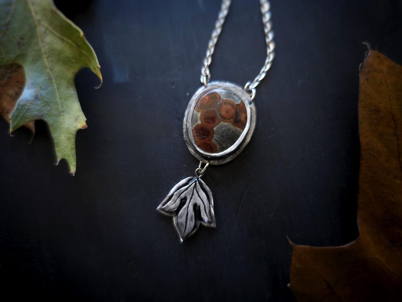May include: Silver pendant necklace with an oval stone featuring a brown and gray pattern. A silver leaf charm hangs below the stone. The necklace is on a dark surface with leaves.