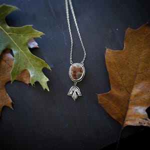 May include: A silver necklace with a pendant featuring a brown and red stone. The pendant is oval-shaped and has a silver leaf design below it. The necklace is displayed with brown and green leaves.