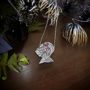 this image shows a hand sculpted knights helmet in silver color. the helmet is decorated with a pink bow