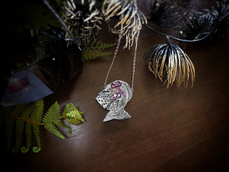 this image shows a hand sculpted knights helmet in silver color. the helmet is decorated with a pink bow