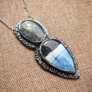 May include: A silver pendant necklace with two connected gemstone pendants. One pendant is oval-shaped with a speckled gray stone, and the other is a teardrop shape with blue, white, and black layers. The necklace is on a brown textured surface.
