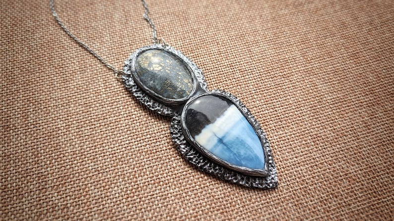 May include: A silver pendant necklace with two connected gemstone pendants. One pendant is oval-shaped with a speckled gray stone, and the other is a teardrop shape with blue, white, and black layers. The necklace is on a brown textured surface.