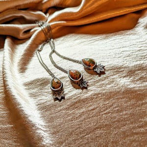 May include: Three silver necklaces with teardrop-shaped pendants featuring a mix of brown, orange, and green colors. Each pendant has a small silver leaf charm.