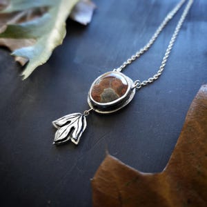 May include: A silver necklace with a pendant featuring a brown and gray stone set in a silver bezel. A silver leaf charm hangs below the stone. The necklace is displayed on a dark surface with autumn leaves.