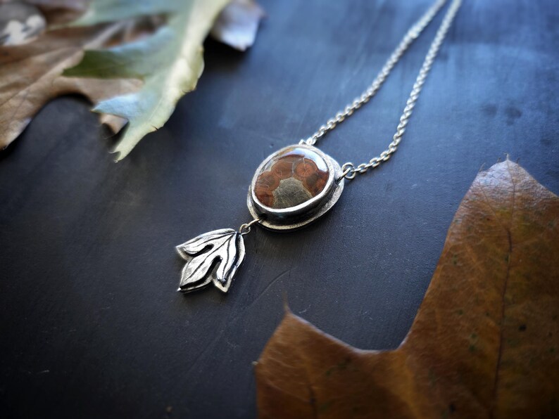 May include: A silver necklace with a pendant featuring a brown and gray stone set in a silver bezel. A silver leaf charm hangs below the stone. The necklace is displayed on a dark surface with autumn leaves.