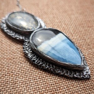 May include: A silver-toned pendant necklace featuring two gemstone settings. The top setting is oval, while the bottom is a teardrop shape with blue and white hues. The pendant is on a silver chain.