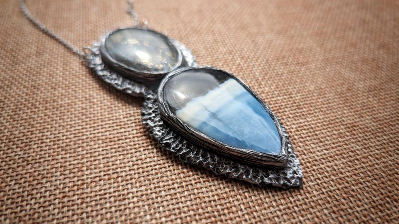 May include: A silver-toned pendant necklace featuring two gemstone settings. The top setting is oval, while the bottom is a teardrop shape with blue and white hues. The pendant is on a silver chain.