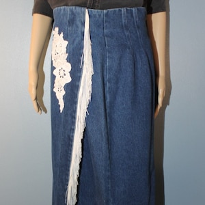 May include: A long denim skirt with a white crocheted flower design on the side. The skirt has a fringe trim around the bottom and down the side.