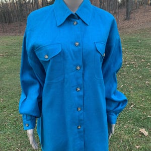 Può includere: Una camicia a maniche lunghe blu turchese con colletto abbottonato e due tasche a patta. La camicia ha bottoni decorativi.