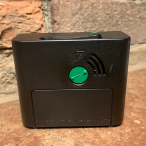 May include: Black alarm clock with a green button and a wireless symbol on the back. The clock has a small battery compartment on the bottom.