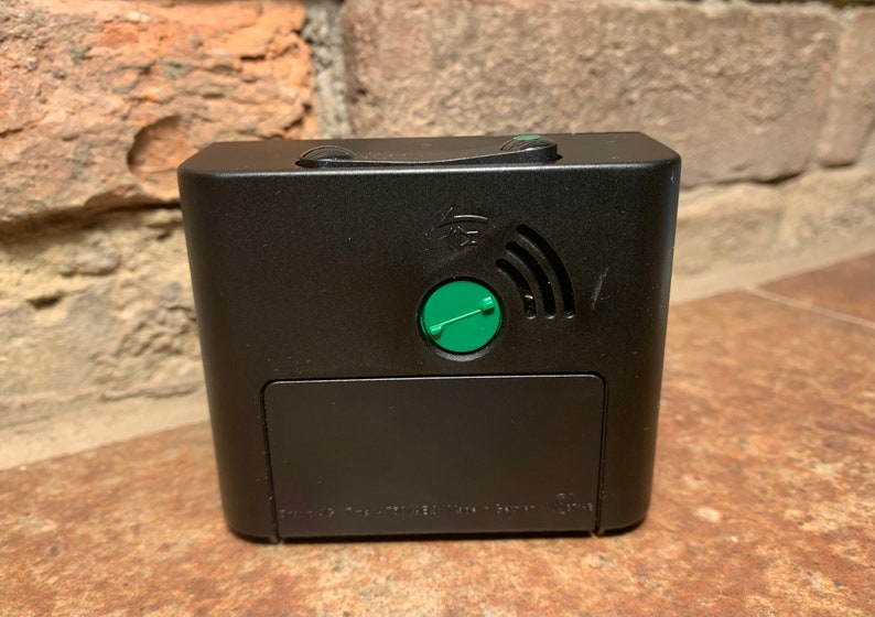 May include: Black alarm clock with a green button and a wireless symbol on the back. The clock has a small battery compartment on the bottom.