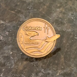 May include: A gold-colored pin with the words "Columbia" and "ENGLE-TRULY" on it. The pin features a bald eagle flying over a stylized American flag with a space shuttle in the background.