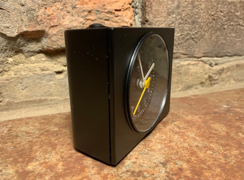 May include: A black alarm clock with a round, black face and white hands. The clock has a yellow second hand and a small, yellow button on the face.