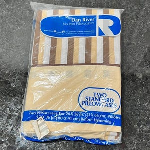 May include: Two standard pillowcases in a plastic package. One set has vertical stripes in brown, tan, and white. The other set is a solid tan color. The packaging reads "Dan River No Iron Pillowcases". Each pillowcase is 20 x 26 inches.