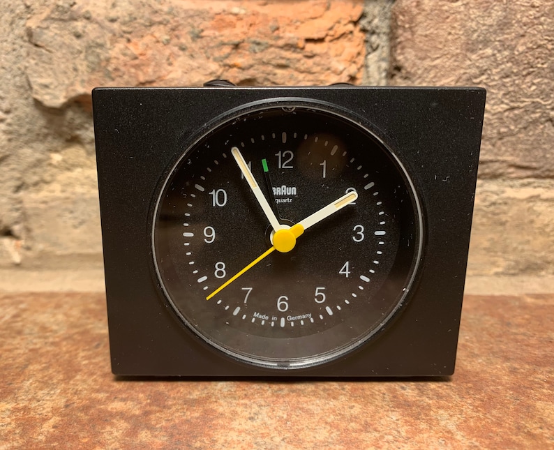 May include: Black alarm clock with a white face and yellow second hand. The clock face has black numbers and the words "Braun quartz" printed on it. The clock is made in Germany.