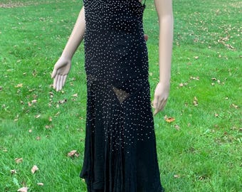 May include: A black beaded evening gown with a plunging neckline and a flowing skirt. The gown is made of a sheer fabric and is adorned with small white beads.