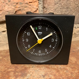 May include: A black Braun quartz alarm clock with a yellow second hand and white hour and minute hands. The clock face is black with white numbers and a white 'Braun quartz' label.