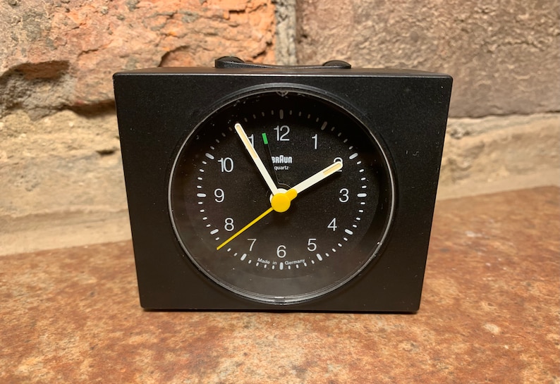 May include: A black Braun quartz alarm clock with a yellow second hand and white hour and minute hands. The clock face is black with white numbers and a white 'Braun quartz' label.