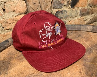 May include: A maroon baseball cap with a flat brim. The cap features embroidered graphics of a football player and a logo, with text that reads "January 29, 1995" and "JOE ROBBIE STADIUM".