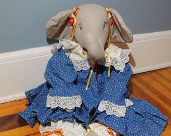 May include: A gray elephant doll wearing a blue dress with white lace trim and yellow ribbons. The dress has a white polka dot pattern. The doll is sitting on a wooden floor.