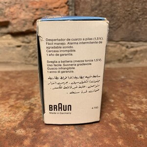 May include: A white box with blue accents for a Braun quartz alarm clock. The box has text in Spanish, Italian, and Arabic describing the clock's features, including a battery-operated alarm, a clear, unbreakable case, and a one-year warranty. The box also has the Braun logo and the text "Made in Germany".
