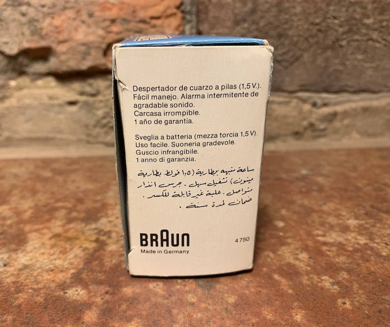 May include: A white box with blue accents for a Braun quartz alarm clock. The box has text in Spanish, Italian, and Arabic describing the clock's features, including a battery-operated alarm, a clear, unbreakable case, and a one-year warranty. The box also has the Braun logo and the text "Made in Germany".